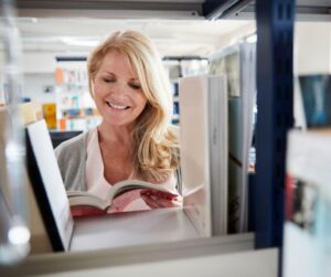 A smiling woman browsing books between library shelves, deciding whether to write fiction or nonfiction.