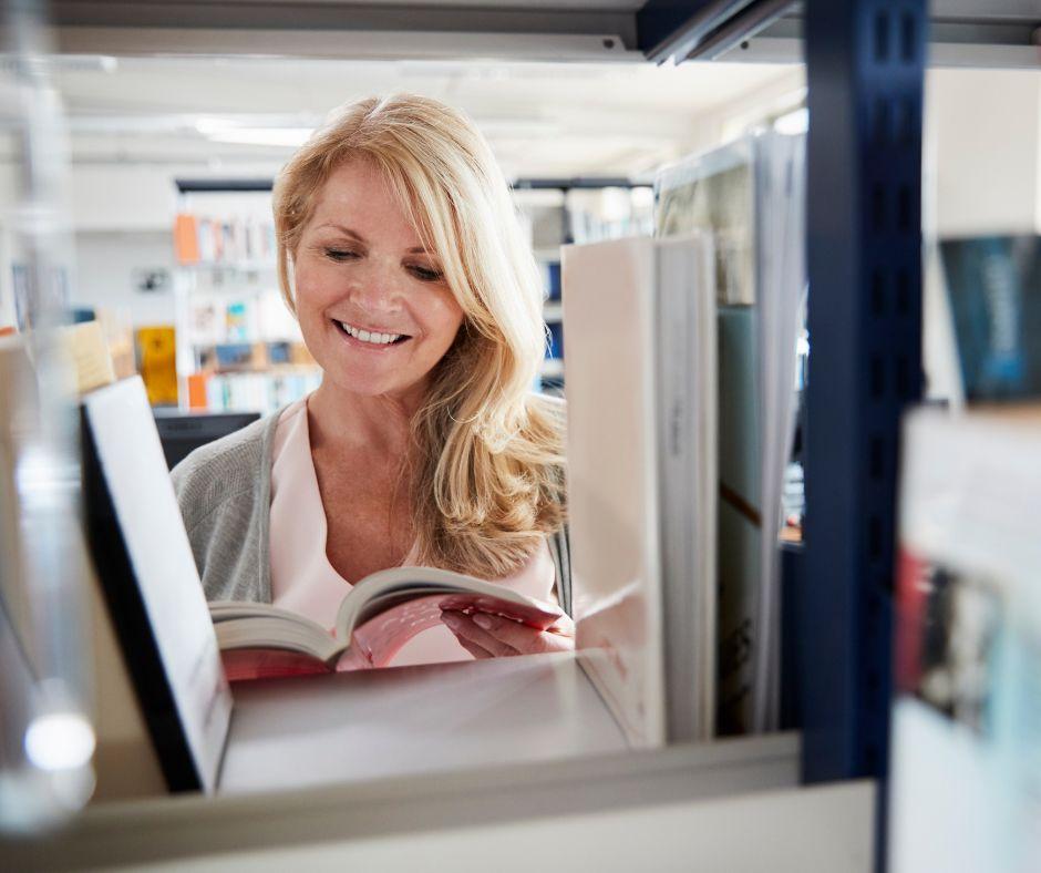 A smiling woman browsing books between library shelves, deciding whether to write fiction or nonfiction.
