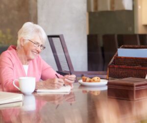 Older woman smiling while writing in a notebook at a table, drafting the opening pages of her memoir