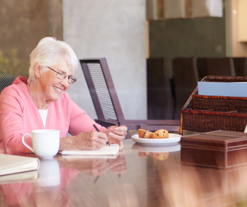 Older woman smiling while writing in a notebook at a table, drafting the opening pages of her memoir