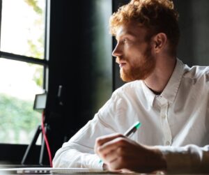 Red-haired man in a white shirt sitting at a desk, thoughtfully looking away while holding a pen over a notebook near his laptop