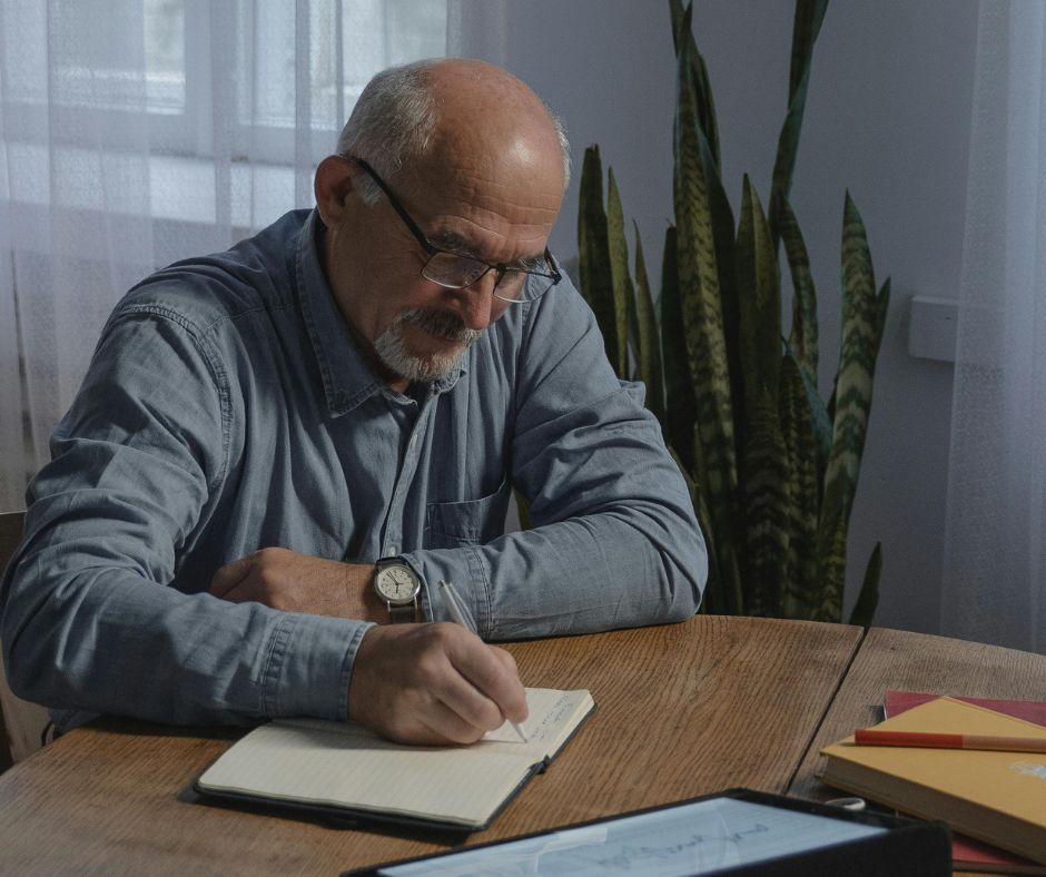Older man in glasses writing by hand in a notebook at a wooden table, surrounded by books and a tablet